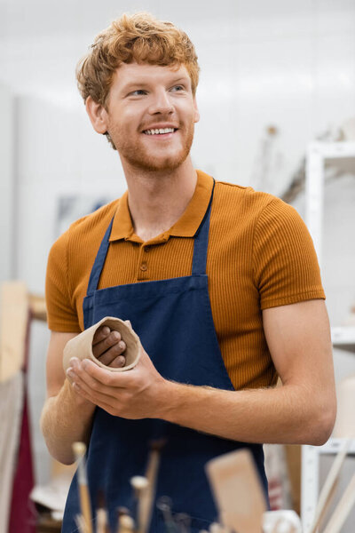 cheerful and bearded man in apron shaping clay piece into cup during pottery class