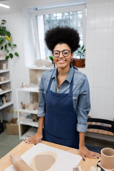 happy young african american woman in eyeglasses and apron standing near rolling pin in pottery workshop
