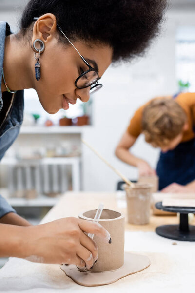 happy african american woman cutting clay around shaped cup near blurred man during pottery class