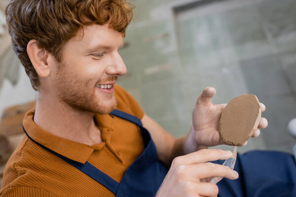 cheerful and redhead man in apron cutting piece of clay with knife