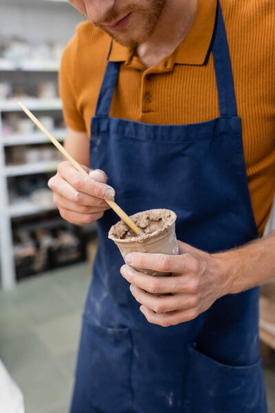 cropped view of bearded man holding paintbrush and clay cup in hands