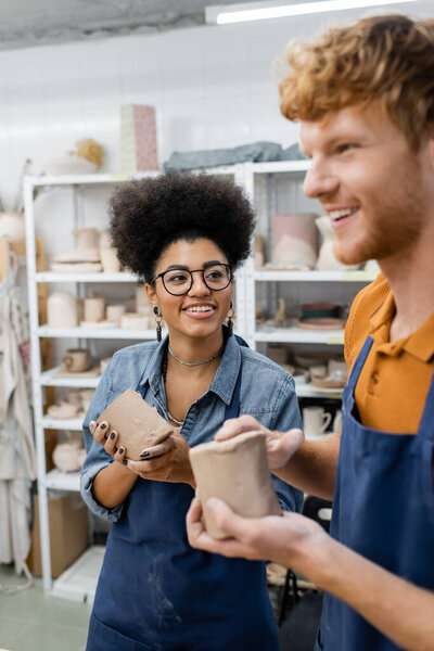 happy african american woman looking at redhead boyfriend molding clay cup in pottery studio 
