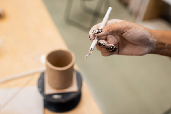 Cropped view of african american craftswoman holding tool near blurred clay in pottery studio 