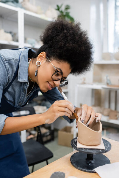 Smiling african american craftswoman in eyeglasses working on shape of clay in pottery studio 