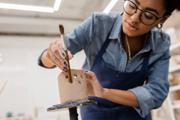 African american sculptor in eyeglasses making holes in ceramic product in workshop 