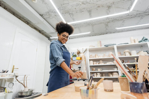 Young african american craftswoman smiling at camera near clay and tools in pottery workshop 