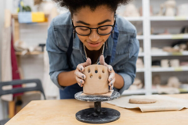 Smiling african american woman in eyeglasses forming clay sculpture in pottery studio 