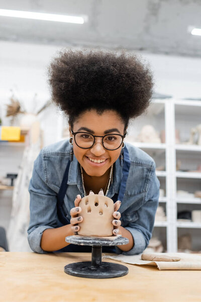 Cheerful african american craftswoman in eyeglasses making ceramic product in pottery workshop 