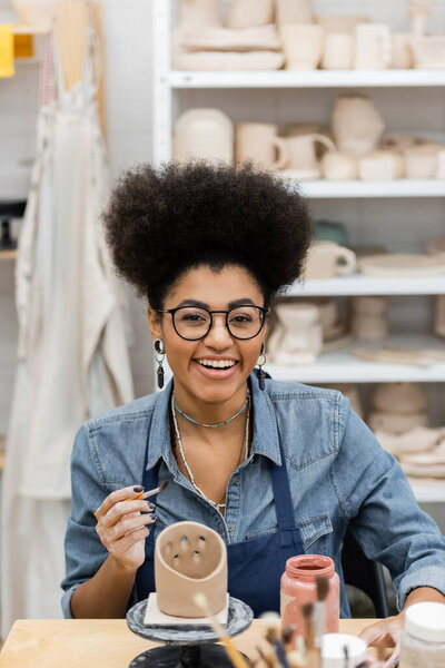 Smiling african american woman in eyeglasses holding paintbrush near clay sculpture and paint in pottery workshop 