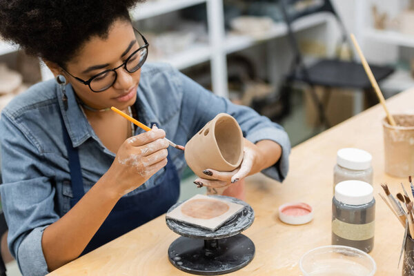 African american artisan in apron painting on ceramic product near jars with paint in pottery studio 