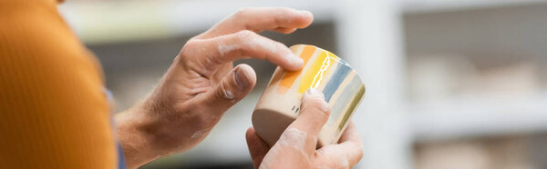 Cropped view of artisan checking ceramic cup in pottery studio, banner 