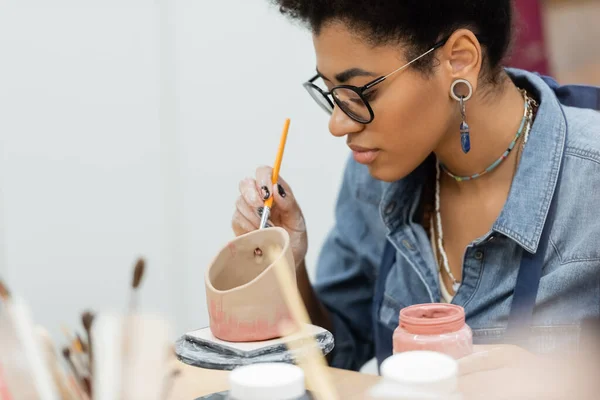 African american artisan in eyeglasses painting on ceramic product near blurred paintbrushes in pottery workshop