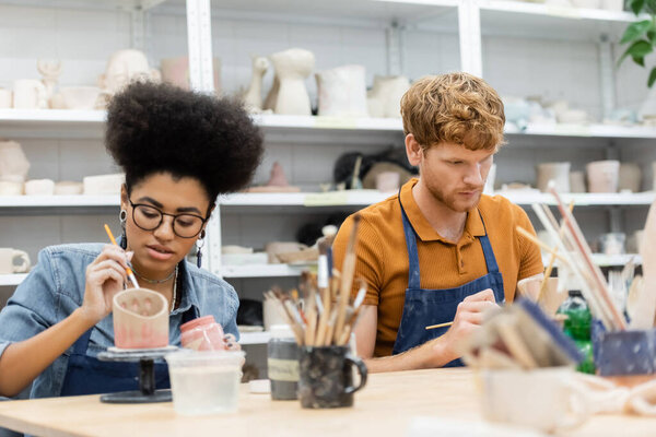 Interracial couple in aprons painting on clay sculptures during date in pottery studio 