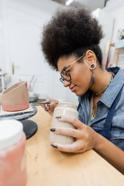 Young african american woman in eyeglasses and apron painting on clay sculpture in pottery studio 