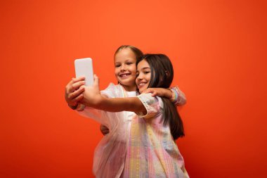 Moda elbiseli neşeli çocuklar portakalda izole edilmiş akıllı telefondan selfie çekiyor ve sarılıyorlar.