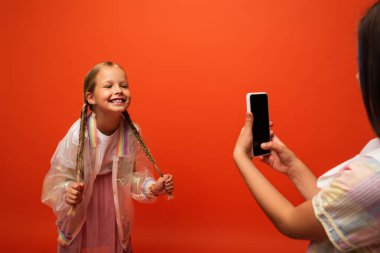 funny girl holding her pigtails near friend taking photo on smartphone isolated on orange clipart