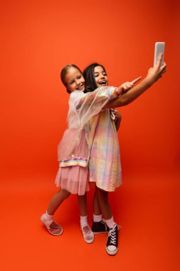 Kızın parmağıyla işaret ettiği boyda. Turuncu arka planda cep telefonundan selfie çekiyor.