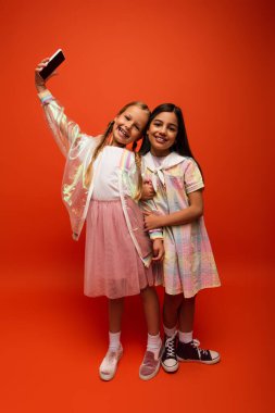 Uzun boylu, yağmurluklu mutlu bir kız, turuncu arka planda bir arkadaşıyla selfie çekiyor.