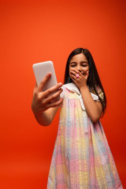Gülen kız elleriyle ağzını kapatıyor ve portakalda izole edilmiş akıllı telefondan selfie çekiyor.
