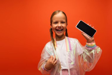 Saçı örgülü heyecanlı bir kız boş ekranlı akıllı telefon gösteriyor ve turuncu üzerine izole bir şekilde gülüyor.