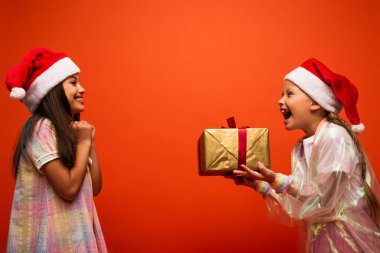 Noel Baba şapkalı hayretler içinde bir kızın elinde Noel hediyesi, turuncu üzerinde izole edilmiş neşeli bir arkadaşın yan görüntüsü.