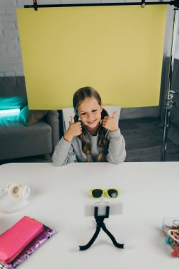 Neşeli çocuk blogcu, sarı arka planda moda güneş gözlüğü ve akıllı telefonun yanında baş parmak gösteriyor.