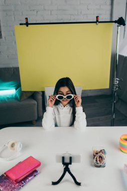 Moda güneş gözlüğü gösteren video blogçusu bulanık akıllı telefonun yanında ve evde fotokopi alanı olan sarı arkaplan