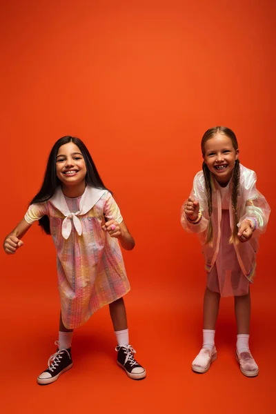 full length of cheerful girls in dress and rain jacket showing clenched fists at camera on orange background