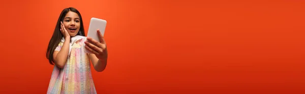 Изумленная Брюнетка Касающаяся Щеки Делающая Селфи Мобильном Телефоне Изолированном Оранжевом — стоковое фото