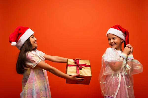 サンタ ハットの笑顔の女の子がオレンジで孤立した幸せな友人にクリスマスプレゼントを — ストック写真