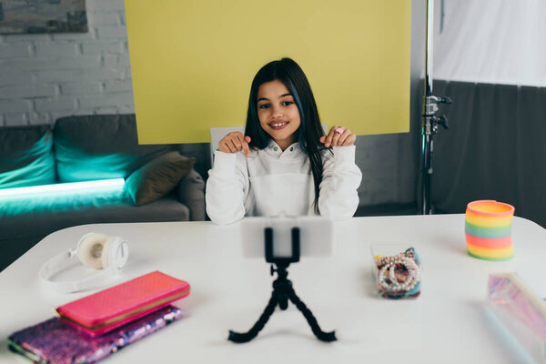 smiling video blogger pointing with fingers at blurred smartphone near pencil cases and beaded bracelets