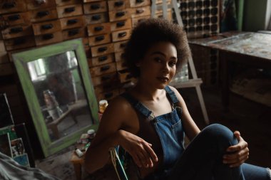 Afro-Amerikan kadın tulumlu, sanat atölyesinde eski ayna ve ahşap mobilyaların yanında oturuyor.