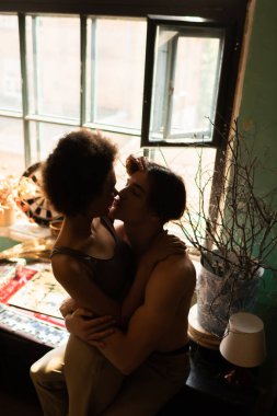 Üstsüz, kaslı bir adamın pencerenin yanındaki Afro-Amerikan kadınını kucaklayıp öptüğü yüksek açılı bir manzara.