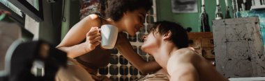 Atölyede birbirlerine bakan seksi gömleksiz adam ve çay bardağıyla Afro-Amerikan kadının yan görüntüsü.