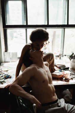 Şehvetli Afrikalı Amerikalı kadın atölyede pencere kenarında oturuyor ve seksi üstsüz erkek arkadaşını öpüyor.