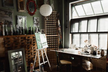 art workshop with large window and vintage furniture near bottles with candles and mirrors on wall  clipart