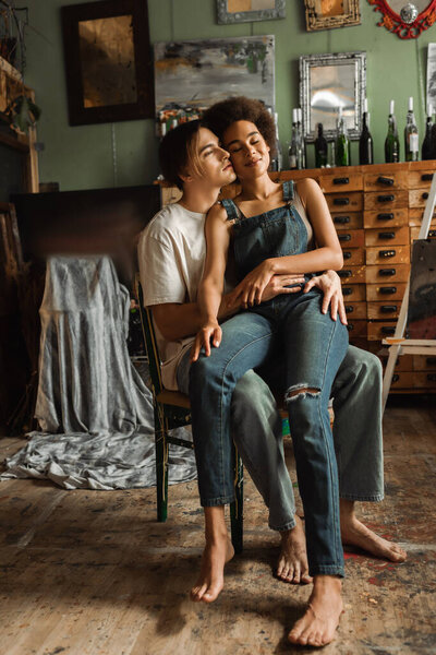young artist hugging sensual african american woman in denim overalls sitting on his laps with closed eyes