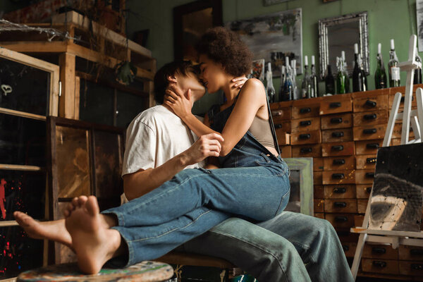 barefoot african american woman in denim overalls kissing with young artist in workshop
