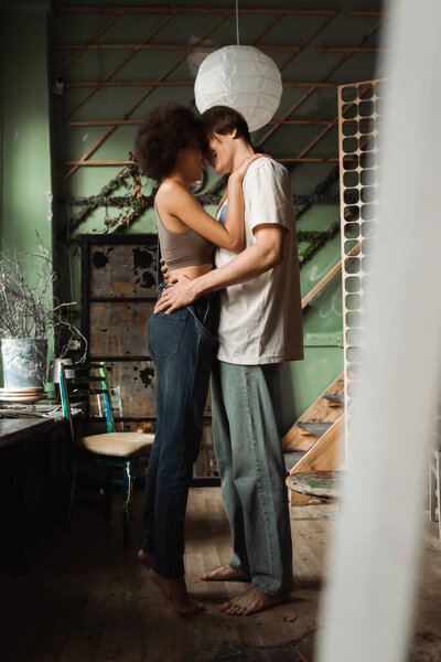 side view of barefoot interracial couple embracing face to face in art studio on blurred foreground