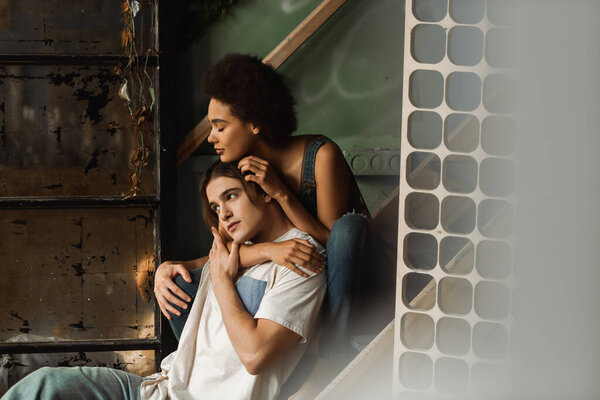pretty african american woman sitting on stairs and hugging pensive boyfriend looking away in workshop