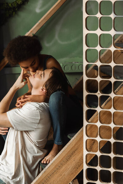 smiling artist touching face of seductive african american woman embracing him on stairs in workshop