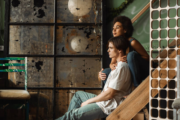dreamy multiethnic couple sitting on stairs near scratched vintage glass and looking away 