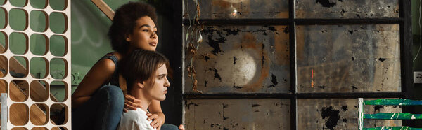 dreamy interracial couple of artists looking away near scratched vintage glass in workshop, banner