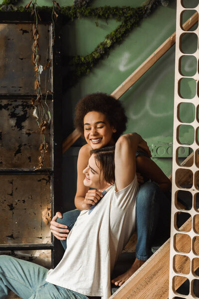 cheerful african american woman with closed eyes embracing with happy man while sitting on stairs in art workshop