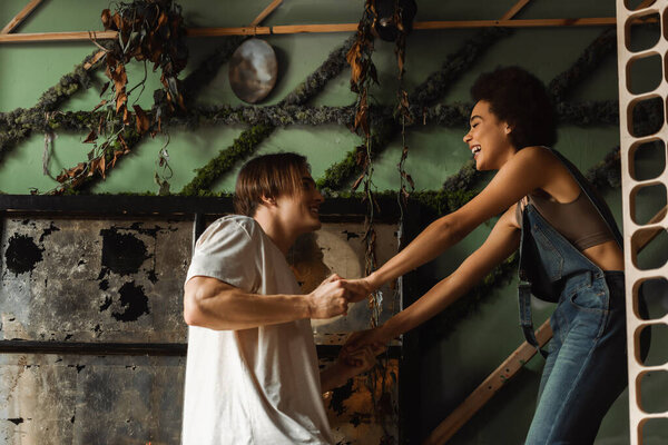 side view of cheerful multiethnic couple holding hands near wall decorated with plants