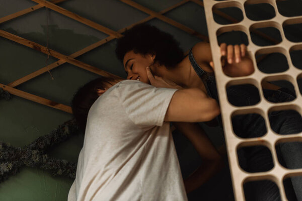 low angle view of man kissing smiling african american girlfriend near blurred decorative lattice in workshop