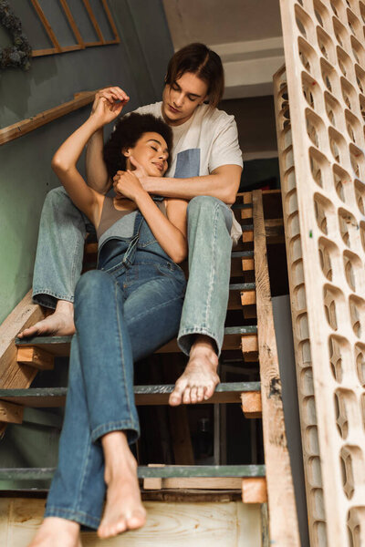 low angle view of barefoot african american woman in denim overalls holding hands with boyfriend on stairs in workshop