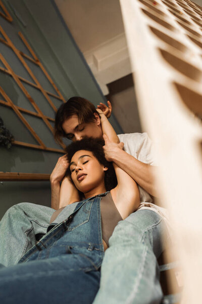 young man with closed eyes holding hands of seductive african american woman in overalls on stairs in workshop