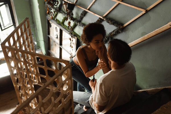 high angle view of young artist sitting on stair in workshop and touching face of sexy african american woman 