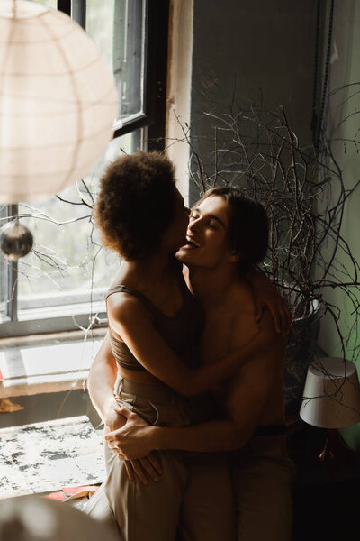 happy shirtless artist hugging young african american girlfriend near window in art studio